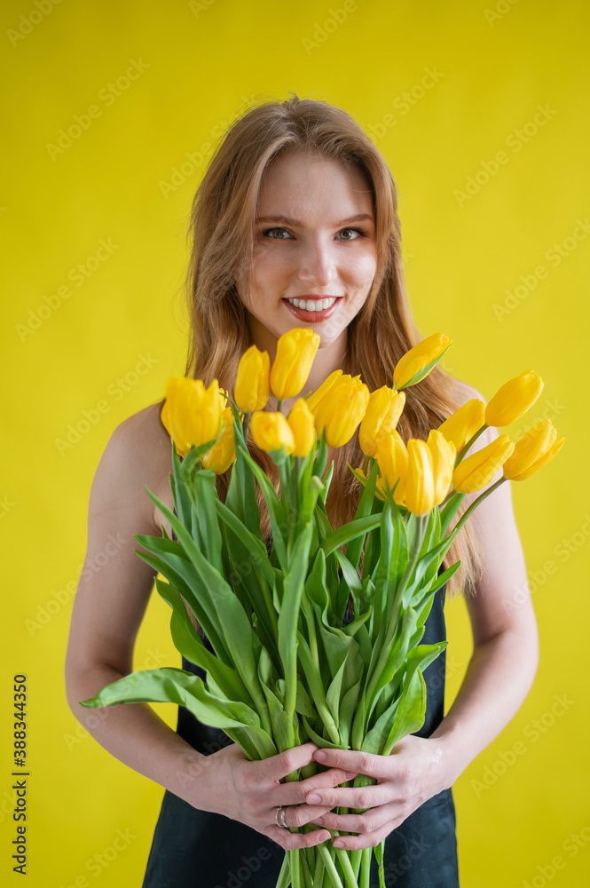 Fototapeta premium Caucasian woman with an armful of yellow tulips on a yellow background. International Women's Day. Bouquet of spring flowers