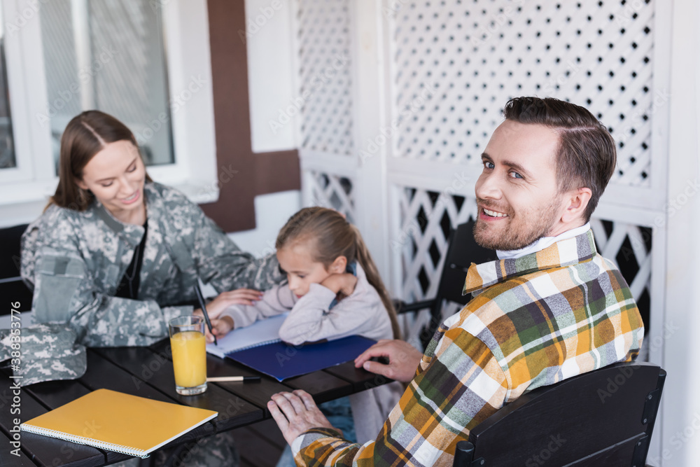 Smiling bearded man in checkered shirt sitting at table with blurred woman and girl on background Smiling bearded man in checkered shirt sitting at table with blurred woman and girl on background