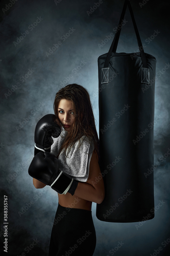 confident sporty woman in black boxing gloves leaning on punching bag on dark studio background with smoke