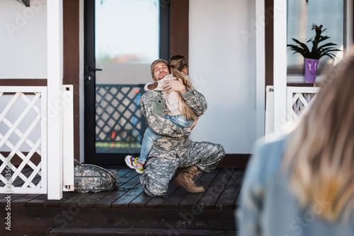 Smiling military service man embracing daughter, while sitting on knee on threshold with blurred woman on foreground