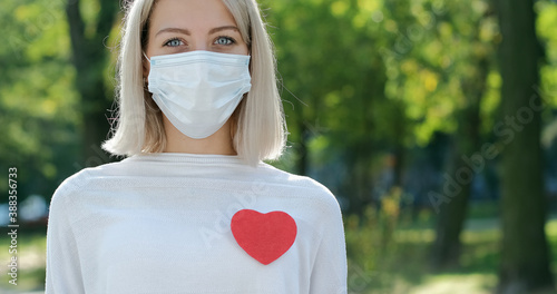 A girl in a medical mask on her face and with a red heart on a jacket stands in the park.