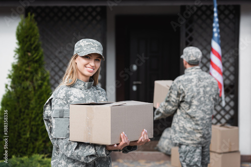Canvastavla Smiling woman in camouflage holding cardboard box and looking at camera with blu