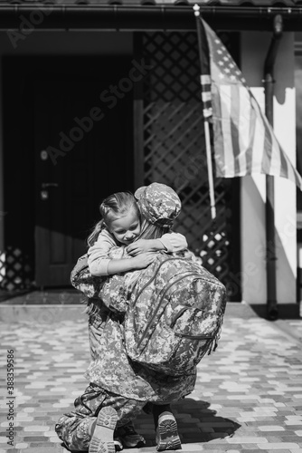 Military servicewoman embracing daughter, while sitting on knee near house and american flag, monochrome