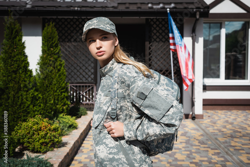 Confident military servicewoman with backpack looking away, while standing near house with american flag