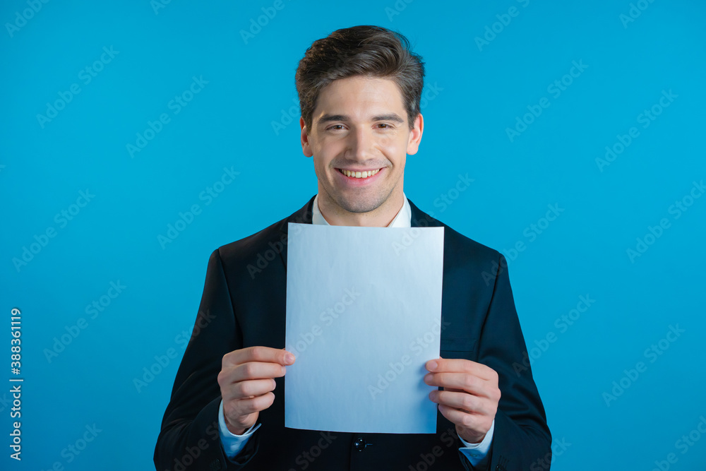 Portrait of young handsome businessman in suit holding white a4 paper isolated on blue studio background.Copy space. Portrait of young handsome businessman in suit holding white a4 paper isolated on blue studio background.Copy space.