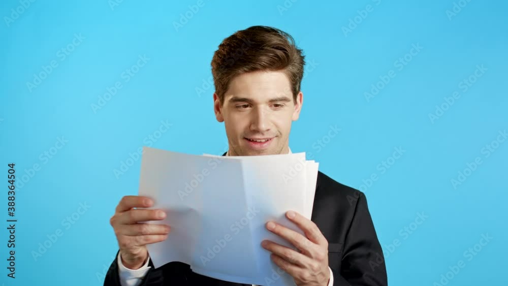 Young handsome man in suit jacket checks documents, utility bills, report. Businessman isolated on blue background. He is satisfied with work of corporate staff. Young handsome man in suit jacket checks documents, utility bills, report. Businessman isolated on blue background. He is satisfied with work of corporate staff.