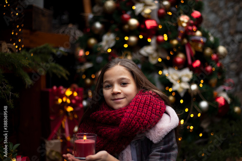 Young pretty pteteen girl  looks at the gift  outdoor at the background decorated christmas tree with lights garland on New Year s Eve holidays. Kid dressed warm jacket  red knitted scarf