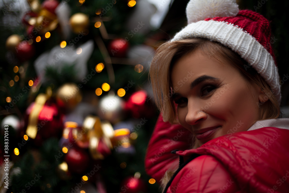 young pretty woman posing outdoor at the background decorated christmas tree with lights garland on New Year's Eve holidays. Kid dressed warm jacket, red knitted hat young pretty woman posing outdoor at the background decorated christmas tree with lights garland on New Year's Eve holidays. Kid dressed warm jacket, red knitted hat