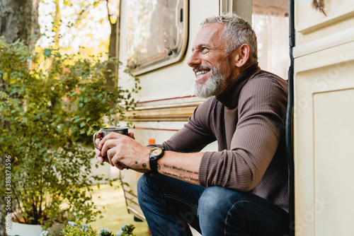 Smiling aged handsome man with beard and tattoo sitting near his trailer holding a mug of coffee