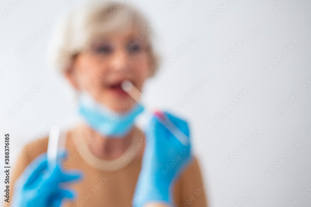 Blurred photo of Testing for presence of coronavirus. Female doctor taking a throat swab test from a senior woman for COVID-19 testing. Doctor testing patient on possible coronavirus infection Blurred photo of Testing for presence of coronavirus. Female doctor taking a throat swab test from a senior woman for COVID-19 testing. Doctor testing patient on possible coronavirus infection