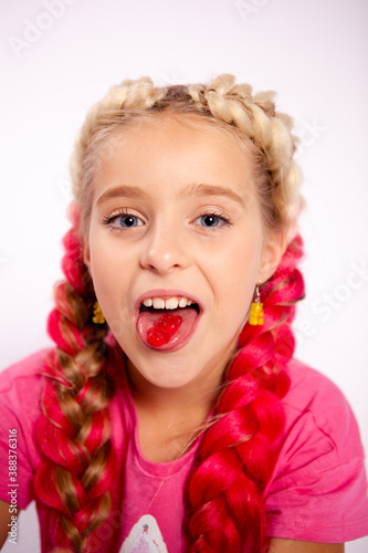 a cute girl in bright colorful clothes and with colored braids shows gelatinous gummies on her tongue