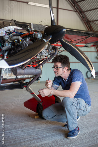 Repairman looking over engine in small aircraft from below. Small red airplane in hangar. Repaiper holding instruments in the hand near the plane engine. Small aviation.