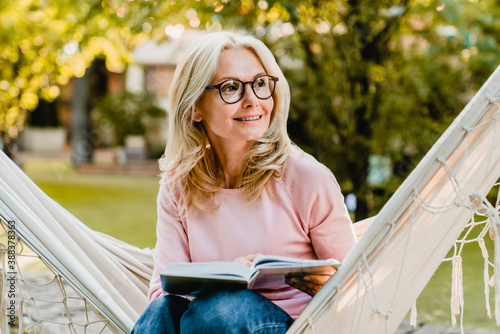 Smiling senior good-looking blond woman wearing glasses while reading in hammock in the summer garden