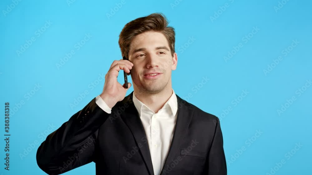 Young businessman speaks with smile on phone. Guy holding and using smart phone. Suit outfit. Blue studio background. Young businessman speaks with smile on phone. Guy holding and using smart phone. Suit outfit. Blue studio background.