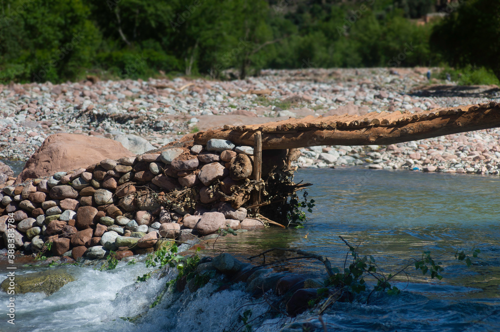 Simple bridge of boulders, pebbles and tree trunks over a fast-flowing river in Setti Fatma, Ourika Valley near Marrakech, Morocco