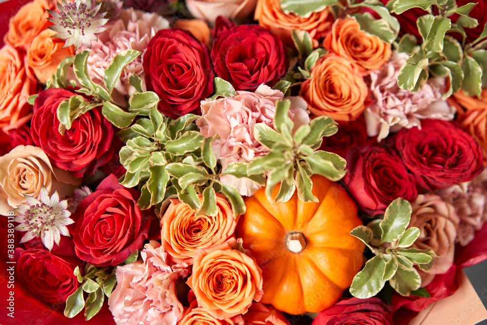 Fototapeta premium Red and orange flowers with pumpkin. Autumn bouquet of mixed flowers in glass vase on wooden table. The work of the florist at a flower shop. Fresh cut flower.