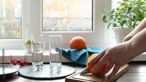 Real woman preparing a blue gin tonic cocktail with ice a mix of alcohol near the window. Cutting orange on wooden board.