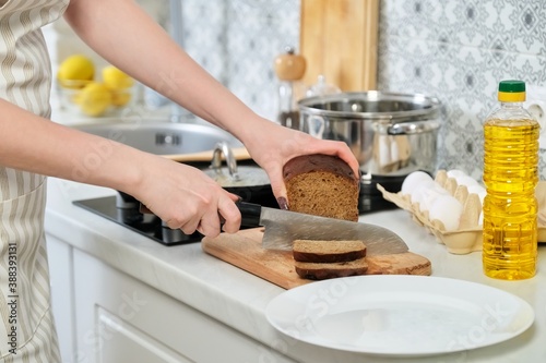 Young woman cuts dark rye bread on cutting board at home in kitchen