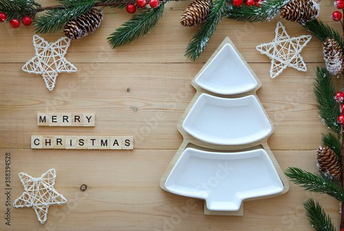 Empty Christmas or new year's plate in white in the shape of a Christmas tree on a wooden background among the branches of a fir tree, decorative stars and the inscription:merry Christmas. Top view. 