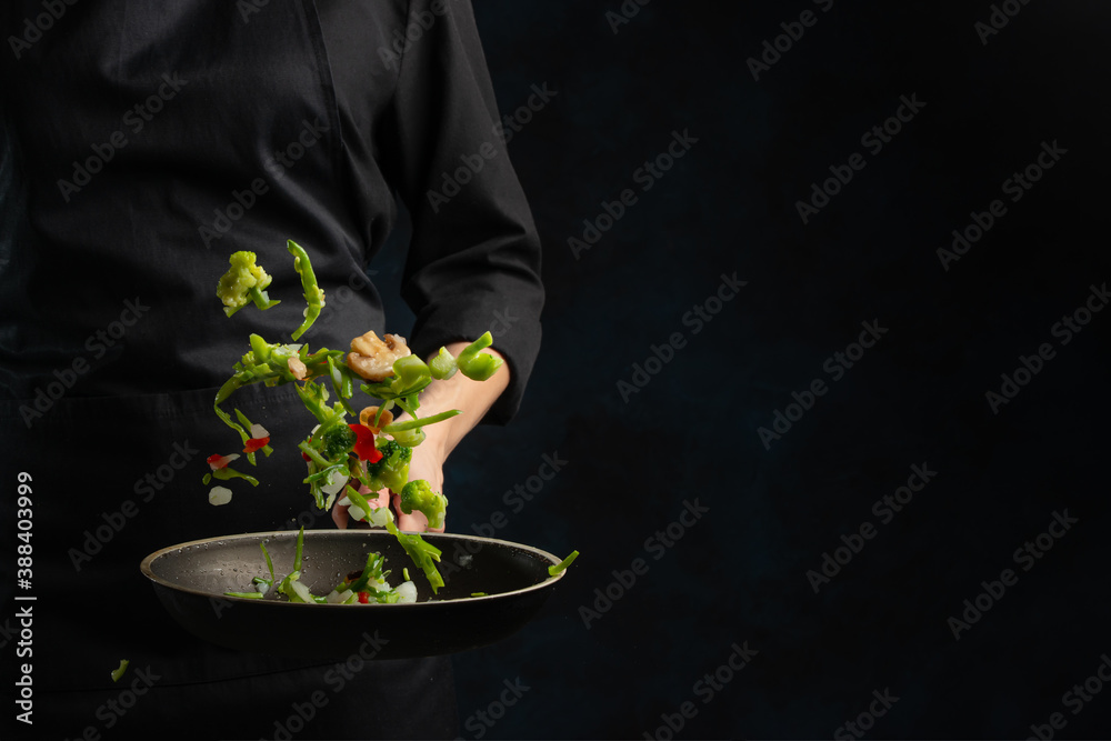 Professional chef in black uniform throws up frying mix of vegetables and mushrooms above the pan on dark blue background. Backstage of cooking meal. Frozen motion. Food banner concept.