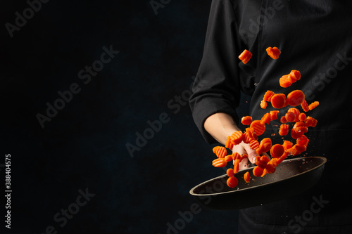 Professional chef in black uniform throws up chopped carrot above the frying pan on dark blue background. Backstage of cooking meal. Frozen motion. Food banner concept.