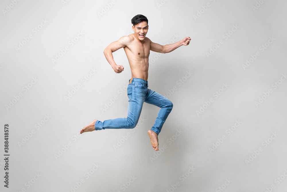 Energetic attractive shirtless Asian man smiling and jumping in light gray studio isolated background