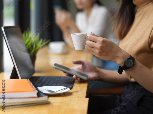 Cropped shot of woman using smartphone while drinking coffee in workspace