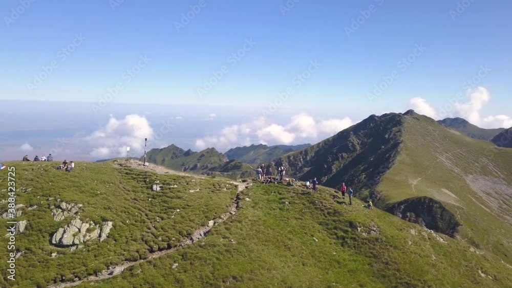 Aerial view of hikers standing on summit of mountain looking out on amazing and dramatic landscape above the clouds. Rugged green terrain of mountain peaks in Romania. Drone orbiting.