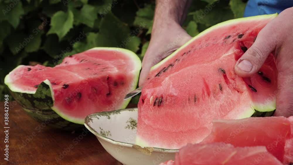 Close-up of a man with a knife cuts off a slice from a watermelon. A large, ripe, striped, green berry lies on the table. We are preparing a dessert for eating at the holiday. Summer day. 4K.