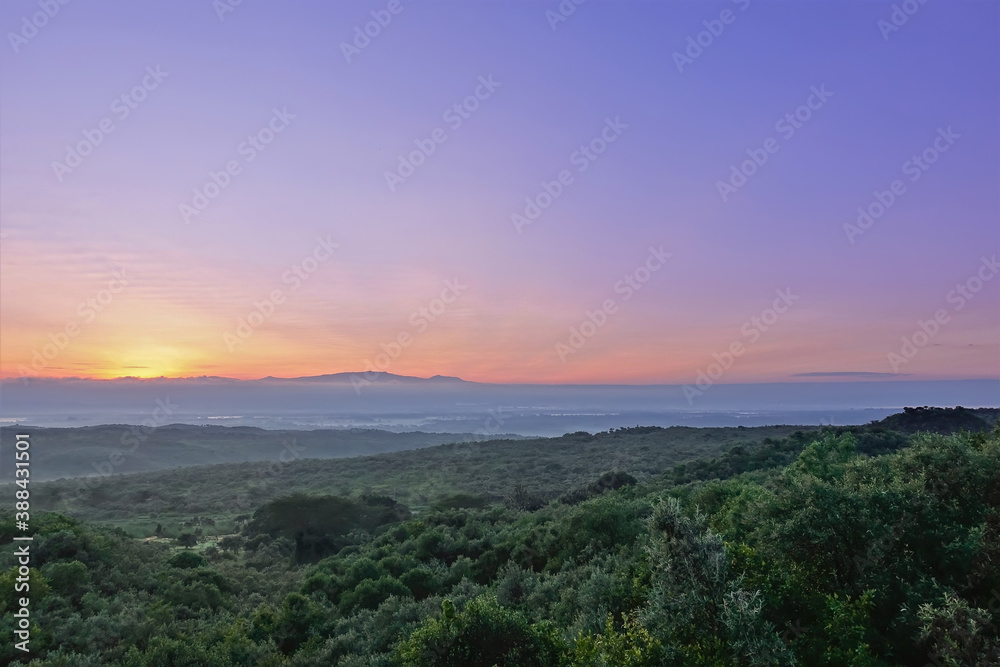 Dawn over the Great Rift Valley. A dense jungle stretches to the horizon. The sun rises from behind the mountains and colors the lilac sky with golden hues. Kenya.