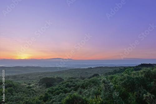 Dawn over the Great Rift Valley. A dense jungle stretches to the horizon. The sun rises from behind the mountains and colors the lilac sky with golden hues. Kenya.