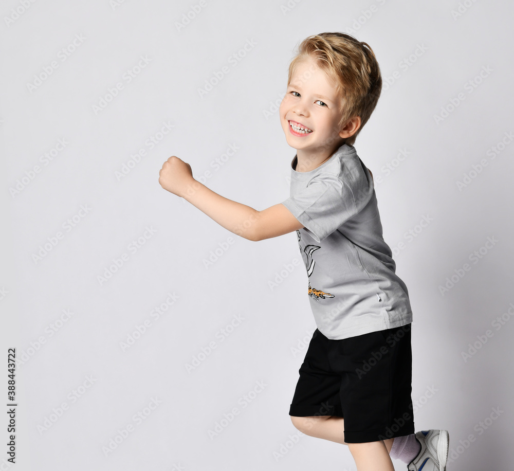 Portrait of cheerful active blond kid boy in grey t-shirt and black shorts running towards copy space. Side view Portrait of cheerful active blond kid boy in grey t-shirt and black shorts running towards copy space. Side view