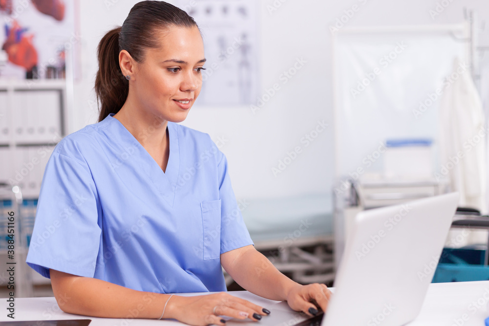 Medical physician typing on laptop sitting at desk wearing blue uniform. Health care physician using computer in modern clinic looking at monitor, medicine, profession, scrubs. Medical physician typing on laptop sitting at desk wearing blue uniform. Health care physician using computer in modern clinic looking at monitor, medicine, profession, scrubs.