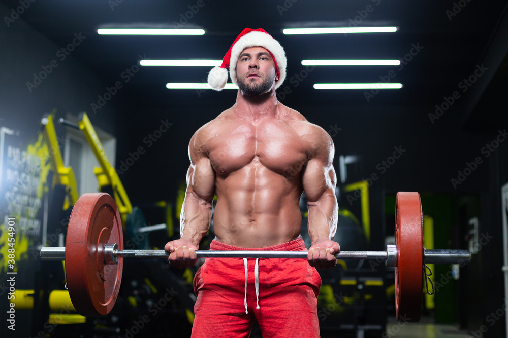 Fototapeta premium Front view of a sexy muscular man wearing Christmas hat and red shorts lifting a barbell in a gym