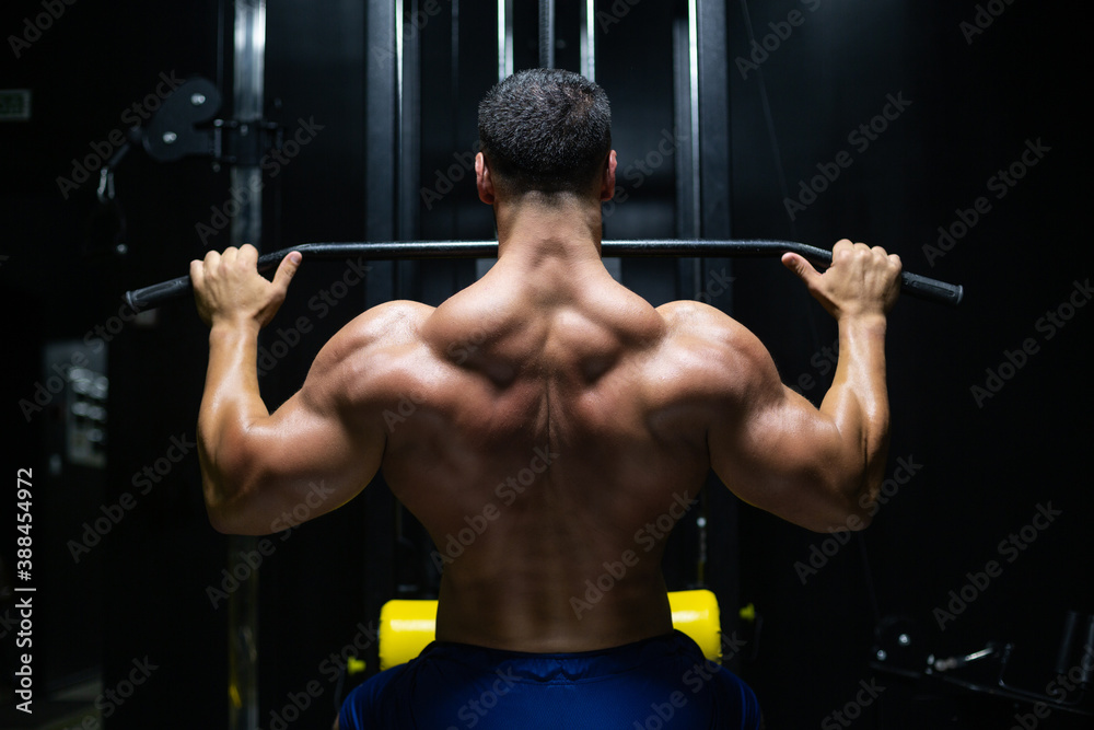 Handsome fitness man is performing back workouts using thrust of the upper block machine in a gym, back view