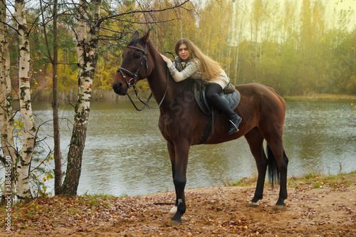 Cute young woman on horseback in autumn forest by lake. Rider female drives her horse in Park in inclement cloudy weather with rain. Concept of outdoor riding, sports and recreation. Copy space © Alex Vog