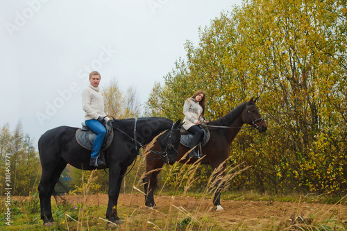 Cute young couple on horsebacks in autumn forest on country road. Riders in autumn Park in inclement cloudy weather with light rain. Concept of outdoor riding, sports and recreation. Copy space © Alex Vog
