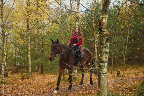 Cute young woman on horseback in autumn forest on country road. Rider female drives her horse in Park inclement cloudy weather with rain. Concept of outdoor riding, sports and recreation. Copy space