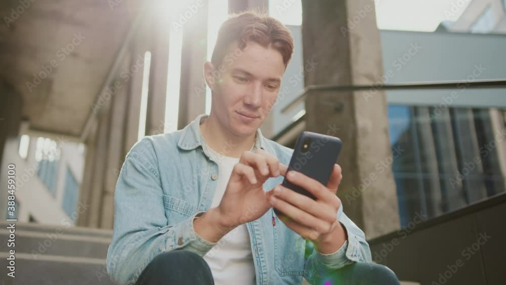 Portrait face student young handsome man with red hair sitting on stairs at sunlight near university or office building. Use smartphone in casual jeans. Slow motion