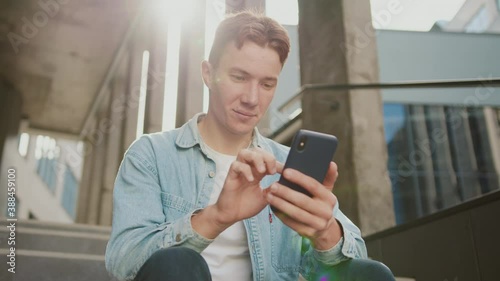 Portrait face student young handsome man with red hair sitting on stairs at sunlight near university or office building. Use smartphone in casual jeans. Slow motion