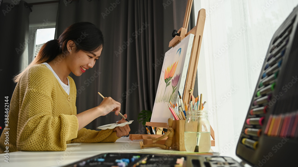 Fototapeta premium Portrait of happy young woman artist holding palette and paintbrush while working on painting in her workshop.