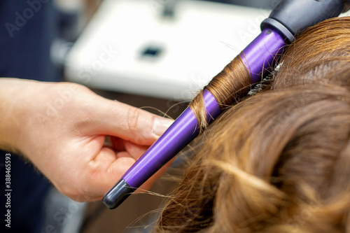 Professional hairdresser makes curly hairstyle by curling iron for long red hair of young woman in hair salon. Soft focus