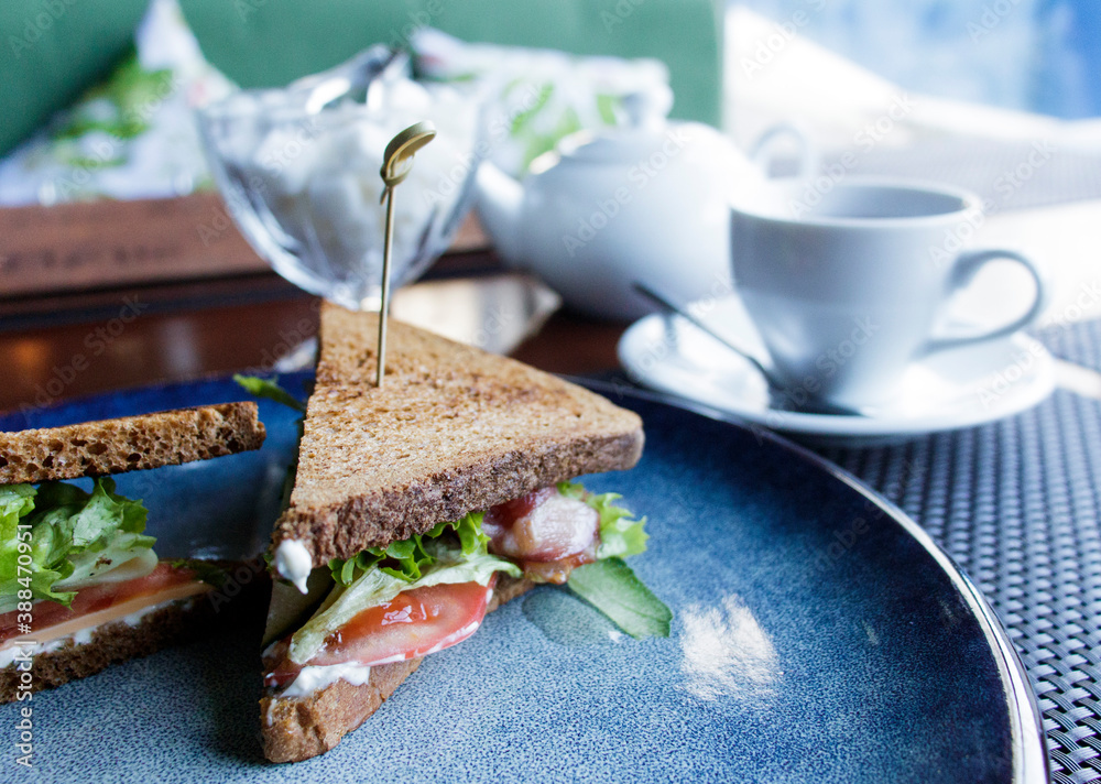 sandwiches with bacon herbs and tomatoes on a dark plate with a white Cup of tea a teapot and a sugar bowl in the background with a blurred background sandwiches with bacon herbs and tomatoes on a dark plate with a white Cup of tea a teapot and a sugar bowl in the background with a blurred background