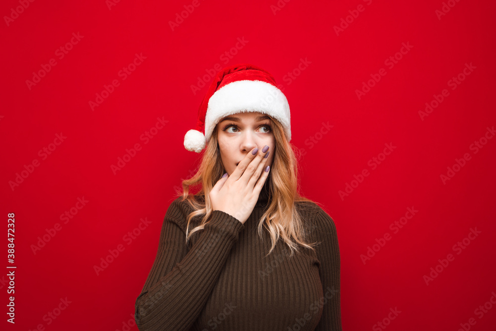 Surprised girl in a warm sweater stands on a red background in a Christmas hat, looks away and covers her mouth with shock. Portrait of shocked girl in santa hat. Surprised girl in a warm sweater stands on a red background in a Christmas hat, looks away and covers her mouth with shock. Portrait of shocked girl in santa hat.
