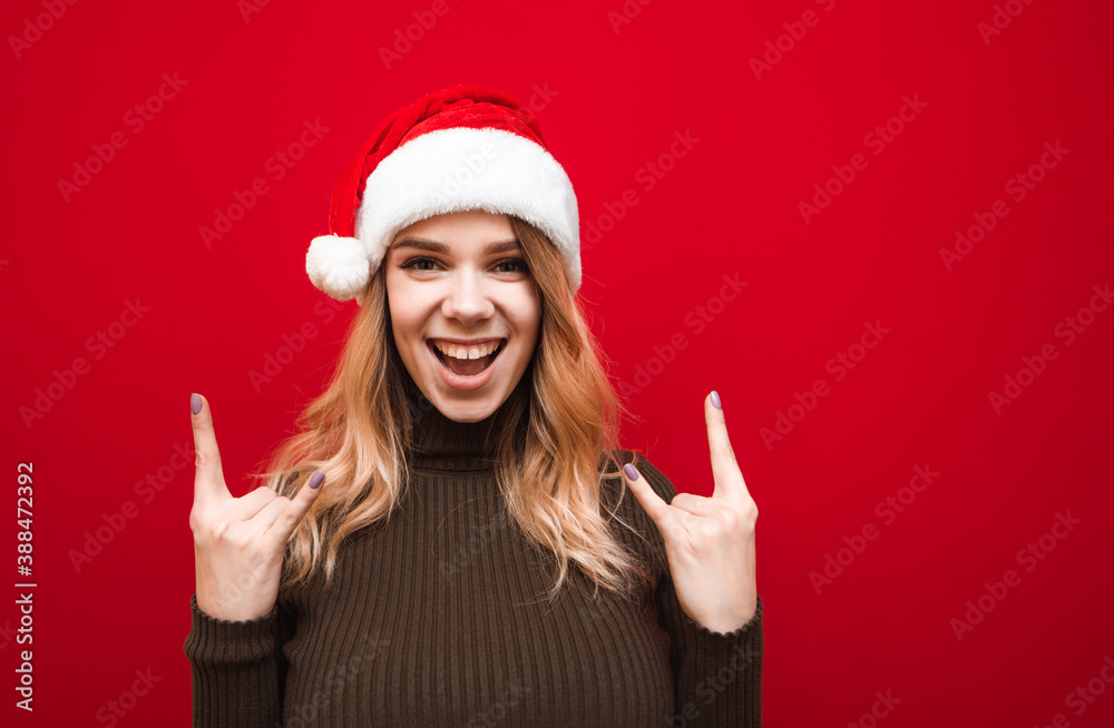 Expressive girl in a warm sweater and Christmas hat stands against a red background, looks at the camera and shows a heavy metal gesture. Isolated. Expressive girl in a warm sweater and Christmas hat stands against a red background, looks at the camera and shows a heavy metal gesture. Isolated.