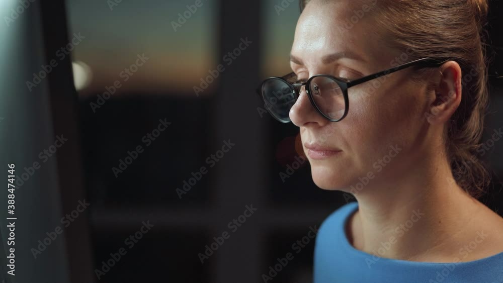 Woman in glasses looking on the monitor and surfing Internet. The monitor screen is reflected in the glasses. Work at night. Home Office. Remote work Woman in glasses looking on the monitor and surfing Internet. The monitor screen is reflected in the glasses. Work at night. Home Office. Remote work