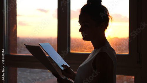 Businesswoman using tablet with sunbeams and lens flare effects against sunset sky window on blurred city background.
