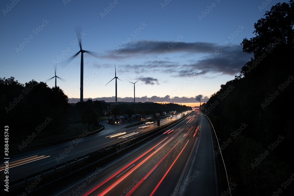 German Autobahn leading past wind turbines towards power plant at sunrise with light trails and motion lblur of the moving cars symbolizing energy and mobility revolution