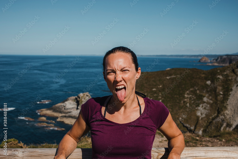 White woman sticking out the tongue, and looking to the camera. Blue sky and ocean in the background.