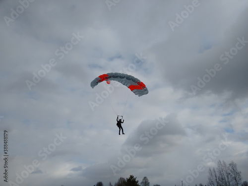 A skydiver lands under a bright multi colored parachute gray blue and orange in winter against a cloudy sky with white and gray clouds